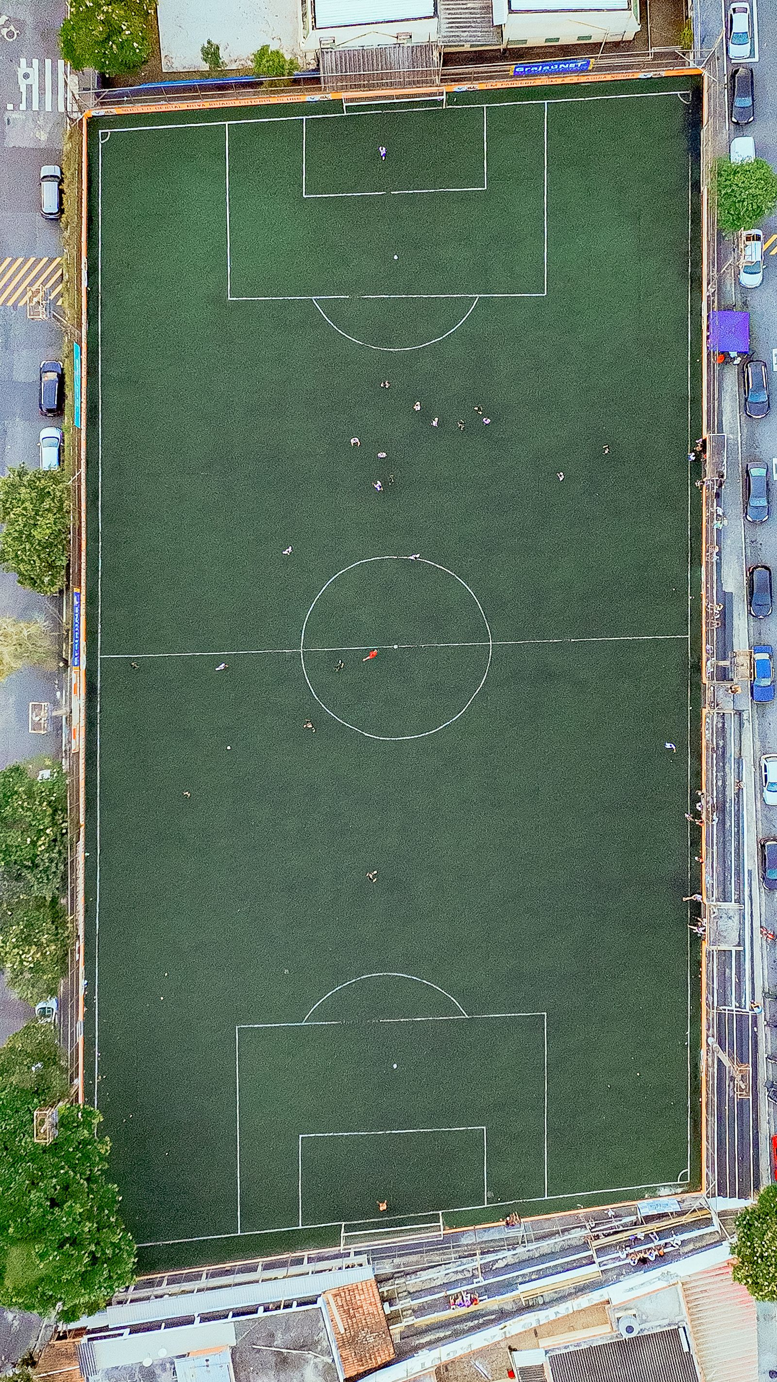 An overhead photograph of a football pitch with players, trees, and surrounding streets